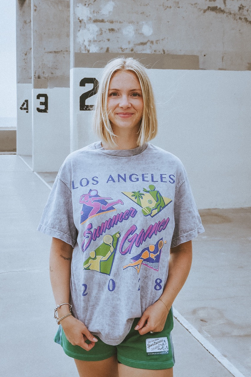 Woman wearing a 'Los Angeles Summer Games 2008' t-shirt standing in front of numbered lockers.