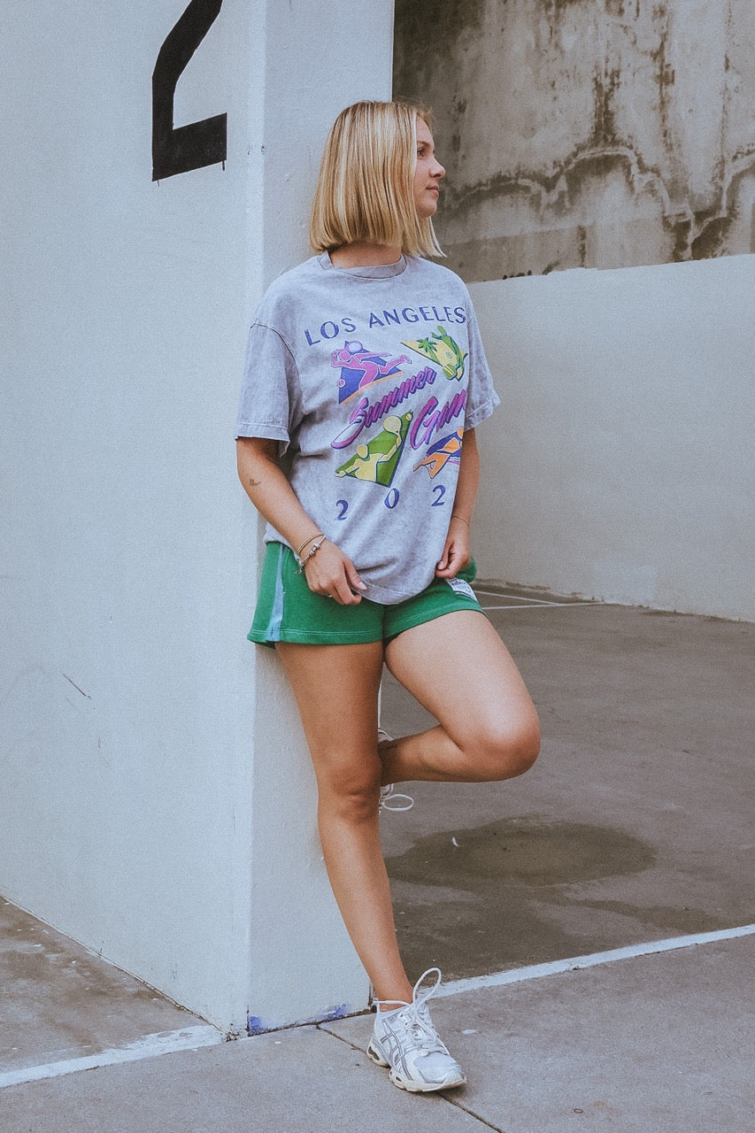 Woman wearing a 'Los Angeles' t-shirt and green shorts leaning against a white wall.